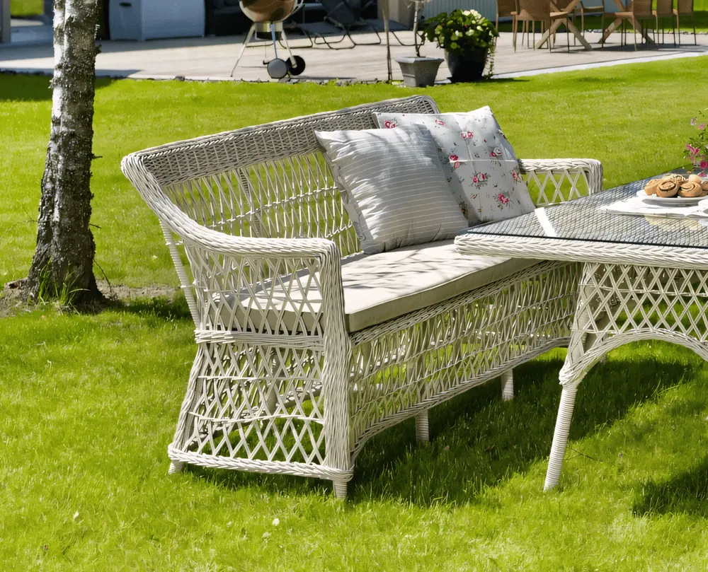 Biała, ażurowa sofa z technorattanu „Orleans” z poduszką na siedzisko i dwiema wzorzystymi poduszkami przy stole na trawniku.