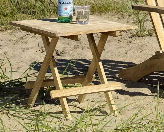 Składany drewniany stolik Bejar z drewna tekowego na piaszczystej plaży, z kieliszkiem i butelką na blacie.