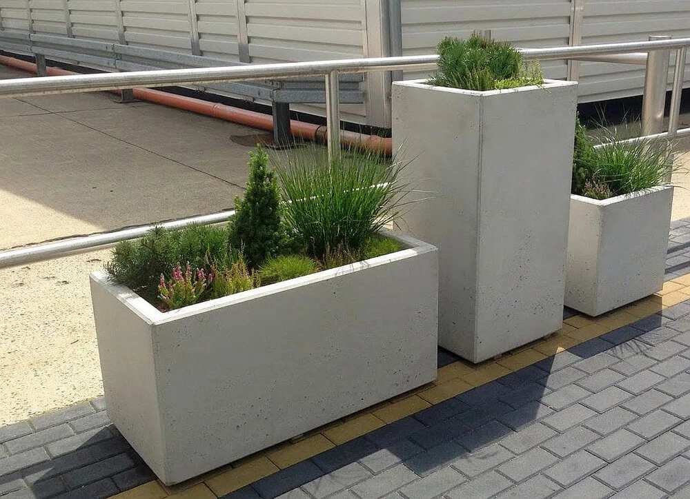 Trzy betonowe donice, w tym betonowa donica o wymiarach 120 x 40 x 50 cm (wys.), z trawami ozdobnymi i krzewami na chodniku.